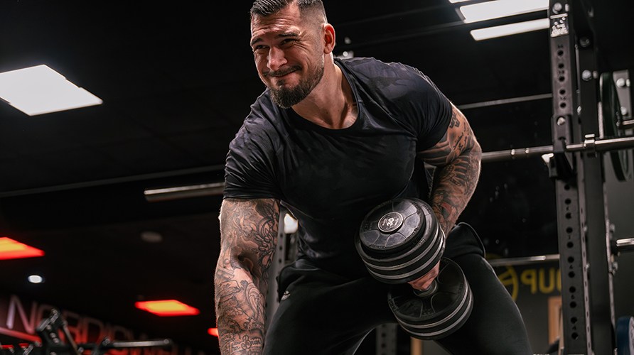 En mand st�r i et tr�ningsmilj� og udf�rer dumbbell row med h�ndv�gte.